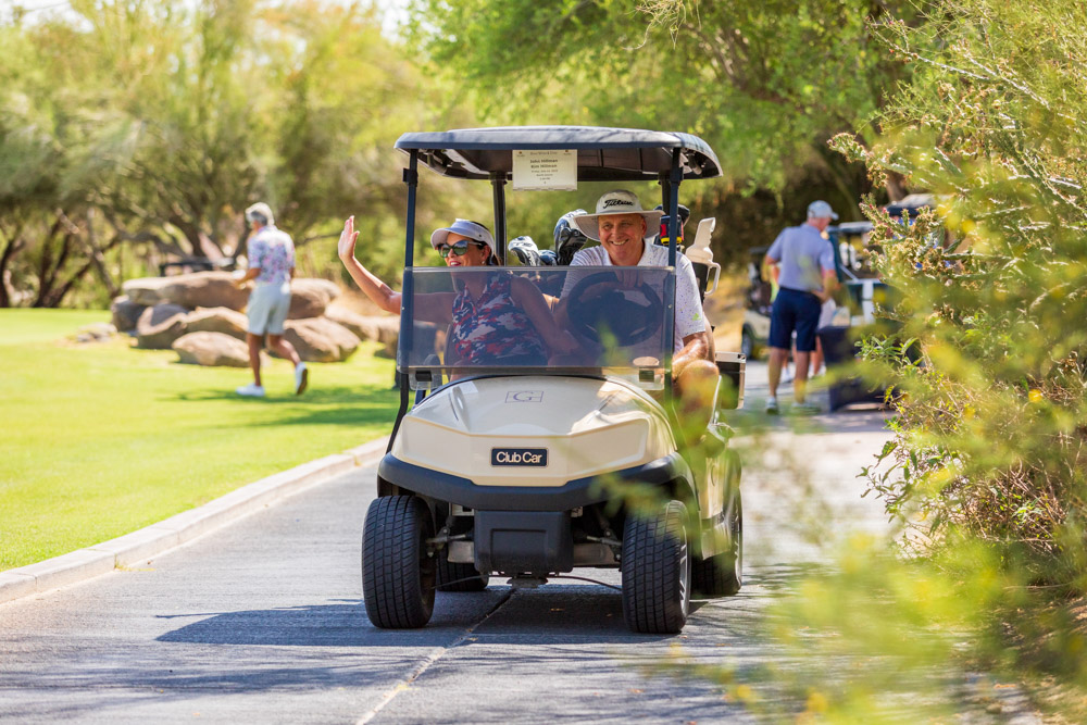 People smiling and waving while riding in a golf cart on a golf course path.