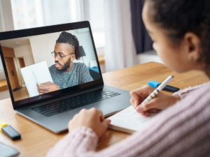 Young girl being tutored online and we can see him on her laptop holding up a piece of paper and pointing to part of it as she takes notes at her home desk.