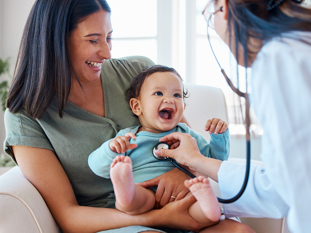 Smiling mom holding smiling baby while Pediatrician checks child with stethoscope.