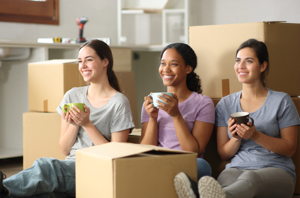 3 female roommates sitting on floor drinking coffee with moving boxes around them.